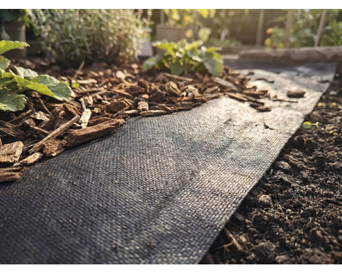 Schwarzes Unkrautvlies in einem Gartenbeet, teilweise mit Rindenmulch bedeckt, neben Erde und kleinen Pflanzen.