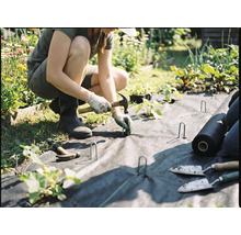 Eine Person verlegt Unkrautvlies im Garten und befestigt es mit Erdankern und einem Hammer.