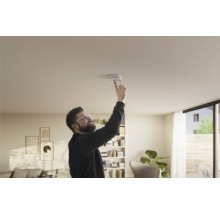 Ein Mann testet einen Rauchmelder an der Decke in einem hell eingerichteten Wohnzimmer.
