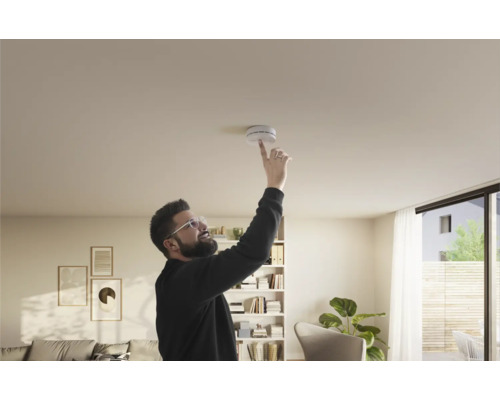 Ein Mann testet einen Rauchmelder an der Decke in einem hell eingerichteten Wohnzimmer.