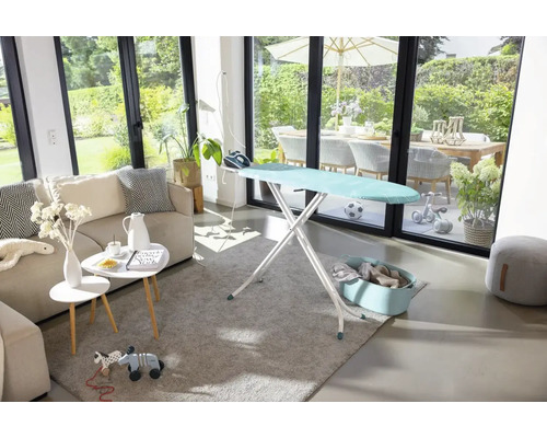 Bügeltisch in einem hellen Wohnzimmer mit Bügeleisen, Wäschekorb, Sofa und Blick auf eine Terrasse.