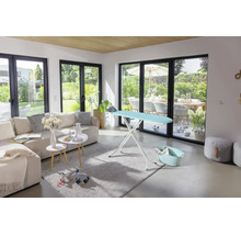 Modernes, helles Wohnzimmer mit großer Fensterfront, einem Bügeltisch auf einem Teppich neben einem Sofa.