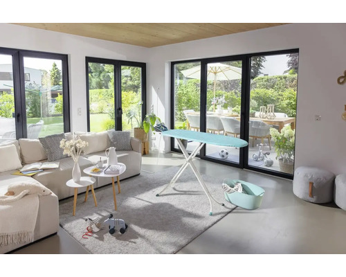 Modernes, helles Wohnzimmer mit großer Fensterfront, einem Bügeltisch auf einem Teppich neben einem Sofa.