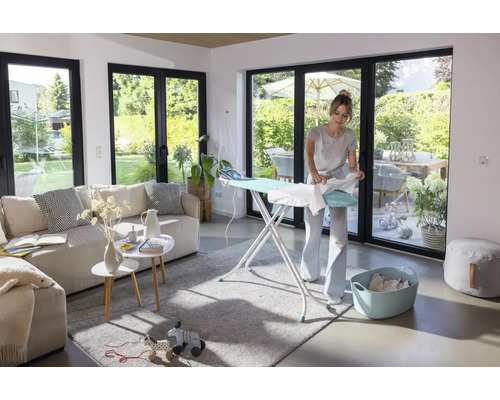 Eine Frau bügelt ein Kleidungsstück auf einem Bügeltisch in einem hellen, modernen Wohnzimmer mit großen Fenstern zum Garten.