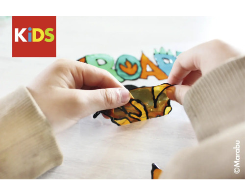 Kinderhände ziehen ein Fensterfarben-Motiv mit Dino-Muster ab. Marabu Logo und KIDS Logo.