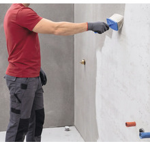 Person trägt mit einem Quast eine Wandgrundierung auf eine graue Wand auf.