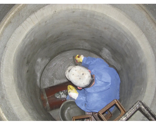 Ein Arbeiter in blauem Schutzanzug dichtet den Boden eines Betonschachts mit Mörtel neben einem Rohr ab.