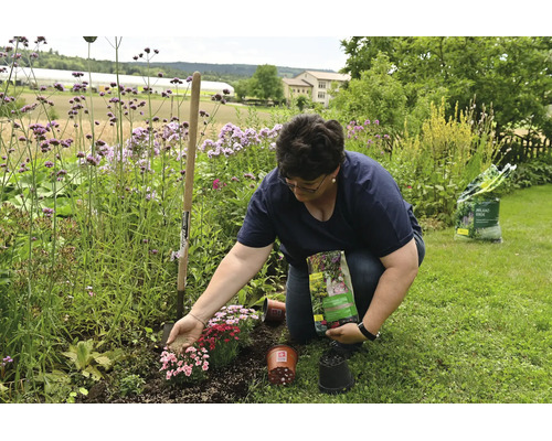 Frau düngt Blumenbeet im Garten mit Dünger, neben ihr stehen ein Spaten, Pflanztöpfe und ein Sack Pflanzerde.