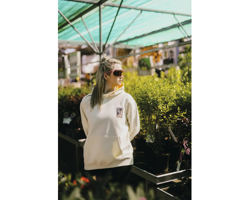 Eine Frau in einem cremefarbenen Hoodie mit Logo steht in einem hellen, organisierten Gartencenter inmitten zahlreicher grüner Pflanzen.