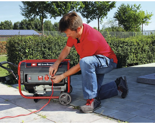 Ein Mann kniet auf einer Terrasse und schließt ein Kabel an einen fahrbaren Einhell Stromerzeuger an.