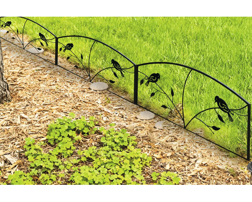 Dekorativer Metall-Gartenzaun mit Vogel- und Ast-Motiv, trennt ein Beet mit Rindenmulch von einer Rasenfläche.