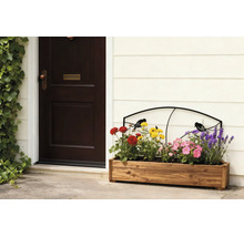 Pflanzkasten aus Holz mit bunten Blumen und einem schwarzen Metallspalier mit Vogelmotiv vor einer hellen Wand neben einer dunklen Haustür.