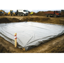 Graue Baufolie flach auf dem Erdboden einer Baustelle ausgelegt mit Baustellenfahrzeugen im Hintergrund.