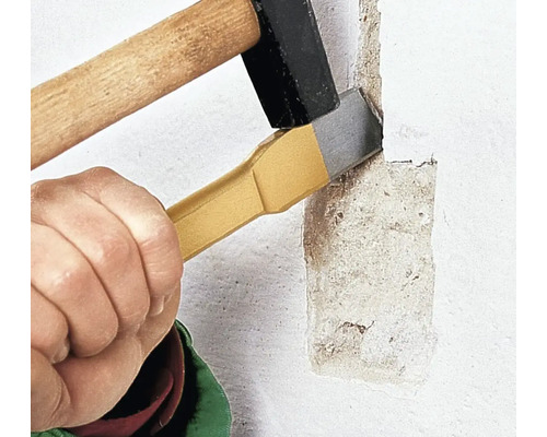 Eine Hand hält einen Flachmeißel an eine verputzte Wand, während ein Hammer auf das Ende des Meißels schlägt, um den Putz abzutragen.