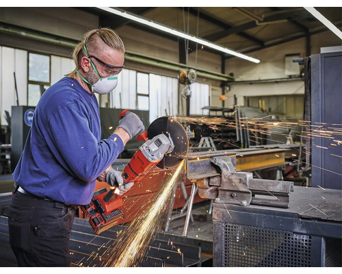 Ein Handwerker in einer Werkstatt nutzt einen Akku-Winkelschleifer zum Trennen eines Metallträgers. Er trägt Schutzbrille, Atemschutz und Handschuhe.