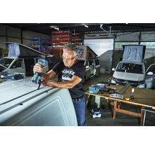 Handwerker nutzt ein Makita Akku-Multitool auf dem Dach eines weißen Kastenwagens in einer Werkstatt mit weiteren Campervans im Hintergrund.