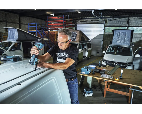 Handwerker nutzt ein Makita Akku-Multitool auf dem Dach eines weißen Kastenwagens in einer Werkstatt mit weiteren Campervans im Hintergrund.