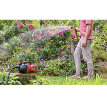 Einhell Akku-Gartenpumpe Aquinna bei der Gartenbewässerung mit einem Gartenschlauch und einer Spritzpistole in einem bepflanzten Garten.