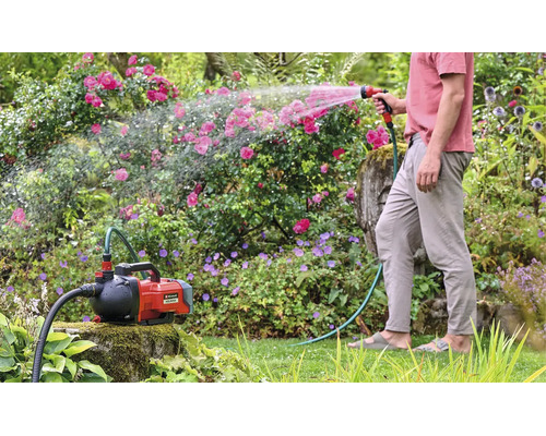 Einhell Akku-Gartenpumpe Aquinna bei der Gartenbewässerung mit einem Gartenschlauch und einer Spritzpistole in einem bepflanzten Garten.