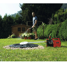 Ein Mann gießt Pflanzen mit einer Einhell Gartenpumpe, die an eine Zisterne im Rasen angeschlossen ist. Ein Gartenhaus steht im Hintergrund.