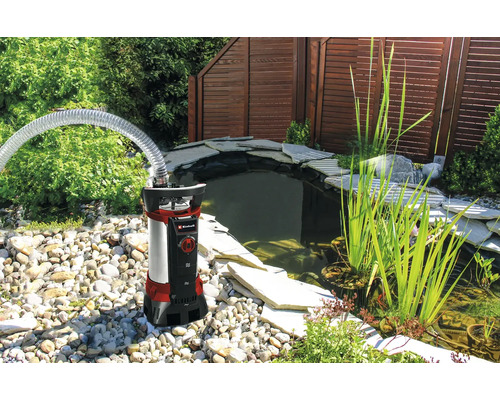 Einhell Tauchpumpe mit transparentem Schlauch an einem Gartenteich mit Steinumrandung und Wasserpflanzen.