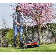 Ein Mann schiebt einen Einhell Akku-Spindelmäher auf einem grünen Rasen im Garten vor einem blühenden Kirschbaum.