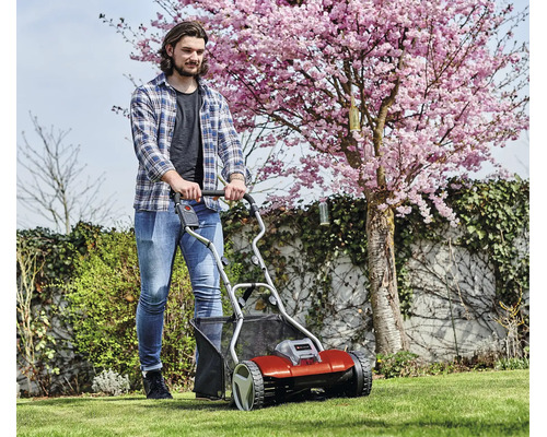 Ein Mann schiebt einen Einhell Akku-Spindelmäher auf einem grünen Rasen im Garten vor einem blühenden Kirschbaum.