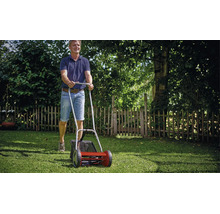 Ein Mann schiebt einen Handspindelmäher mit Grasfangkorb über einen grünen Rasen im Garten.