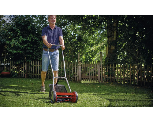 Ein Mann schiebt einen Handspindelmäher mit Grasfangkorb über einen grünen Rasen im Garten.