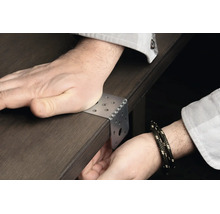 Ein Winkelverbinder aus Metall wird an einer dunklen Holzkante angesetzt und mit der Hand auf einer Tischplatte fixiert.