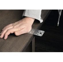 Eine Hand liegt auf einem rechteckigen Lochblech Verbinder aus Metall auf einer Holzoberfläche.