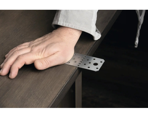 Eine Hand liegt auf einem rechteckigen Lochblech Verbinder aus Metall auf einer Holzoberfläche.