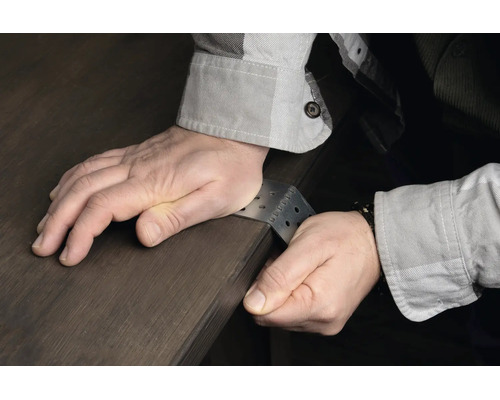 Person biegt einen gelochten Metallwinkel aus Stahl per Hand über eine Holzkante.