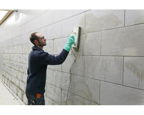 Handwerker beim Verfugen einer gefliesten Wand mit einem Fugenbrett und Schutzhandschuhen