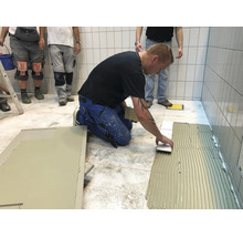 Handwerker beim Verlegen von Bodenfliesen auf einem Fliesenkleberbett in einem gefliesten Raum.