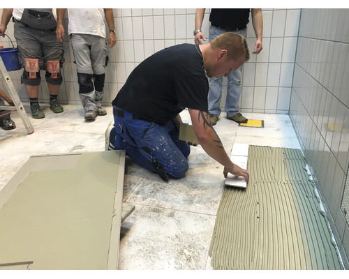 Handwerker beim Verlegen von Bodenfliesen auf einem Fliesenkleberbett in einem gefliesten Raum.