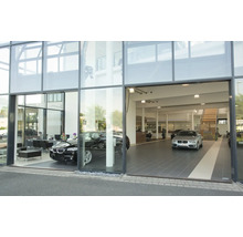 Moderne Glasfassade eines Autohauses mit zwei ausgestellten Fahrzeugen und einer Treppe im Innenraum.