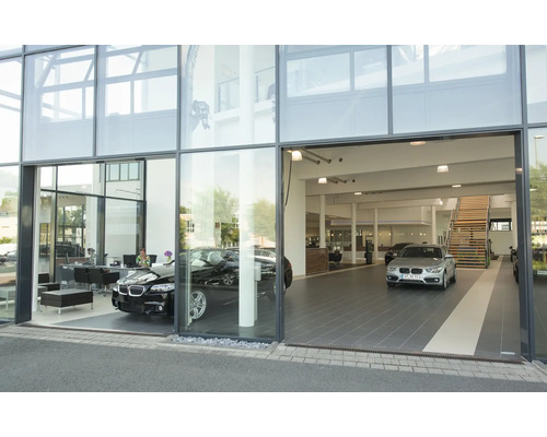 Moderne Glasfassade eines Autohauses mit zwei ausgestellten Fahrzeugen und einer Treppe im Innenraum.
