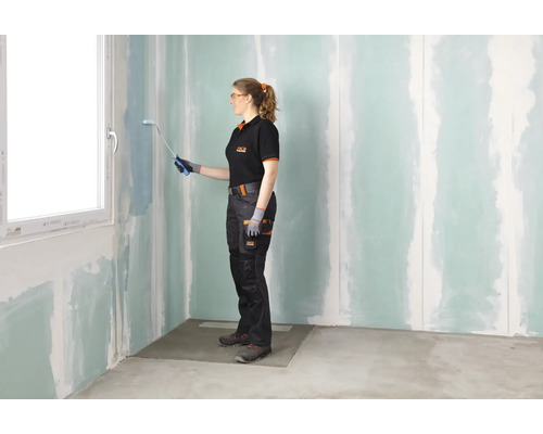 Frau in Arbeitskleidung streicht eine mit Grundierung behandelte Gipskartonwand in einem renovierungsbedürftigen Raum mit einer Farbrolle.