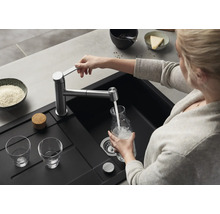 Eine Person füllt eine Glaskaraffe mit Wasser aus einer modernen Küchenarmatur über einem schwarzen Einbauspülbecken in einer Küche.