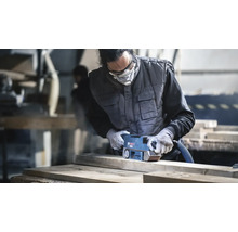 Handwerker schleift ein Holzbrett mit einem Bosch Bandschleifer in einer Werkstatt mit Schutzkleidung und Atemschutzmaske.