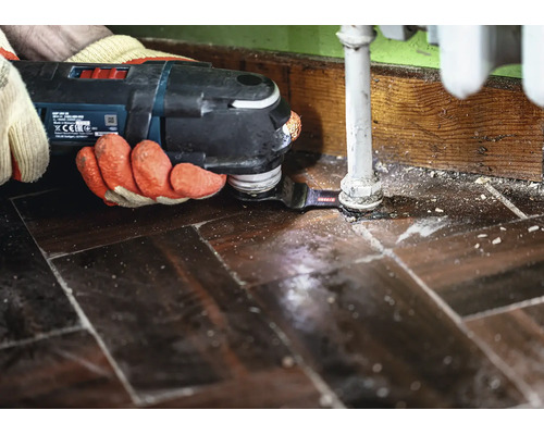 Ein Handwerker nutzt ein Bosch Multifunktionswerkzeug mit Sägeblatt, um den Holzboden passgenau um ein Heizungsrohr herum zu sägen.