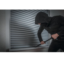Ein Einbrecher versucht mit einem Brecheisen einen geschlossenen Rollladen an einem Fenster aufzuhebeln.
