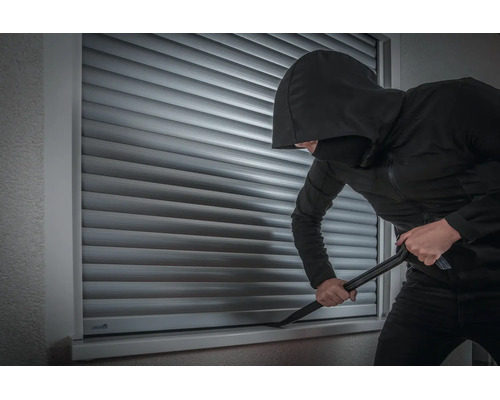 Ein Einbrecher versucht mit einem Brecheisen einen geschlossenen Rollladen an einem Fenster aufzuhebeln.