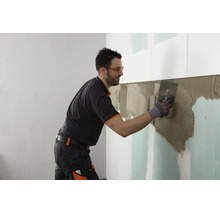 Handwerker beim Auftragen von Fliesenkleber mit einer Zahnkelle auf eine Gipskartonwand bei der Fliesenverlegung.
