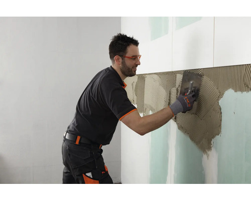 Handwerker beim Auftragen von Fliesenkleber mit einer Zahnkelle auf eine Gipskartonwand bei der Fliesenverlegung.