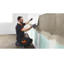 Ein Handwerker in Arbeitskleidung und Schutzbrille bringt eine Fliese auf die mit Fliesenkleber bestrichene Wand auf.