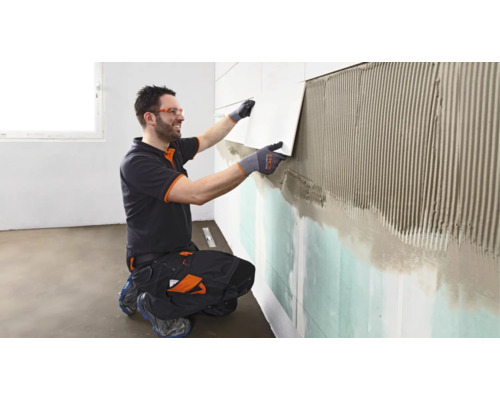 Ein Handwerker in Arbeitskleidung und Schutzbrille bringt eine Fliese auf die mit Fliesenkleber bestrichene Wand auf.
