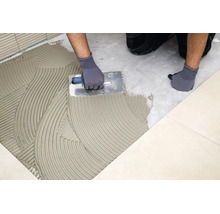 Handwerker beim Auftragen von Fliesenkleber auf den Boden mit einer Zahnkelle zum anschließenden Verlegen von Fliesen.