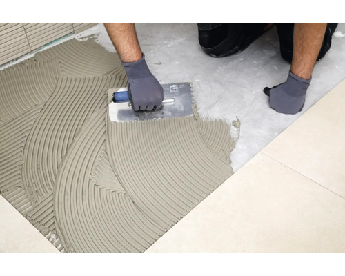 Handwerker beim Auftragen von Fliesenkleber auf den Boden mit einer Zahnkelle zum anschließenden Verlegen von Fliesen.